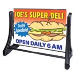 An outdoor advertising sign for "Joe's Super Deli" featuring a large sandwich illustration. The sign highlights "Daily Specials!" in a blue starburst and states "OPEN DAILY 6 AM" at the bottom. The sign is held in a black frame.