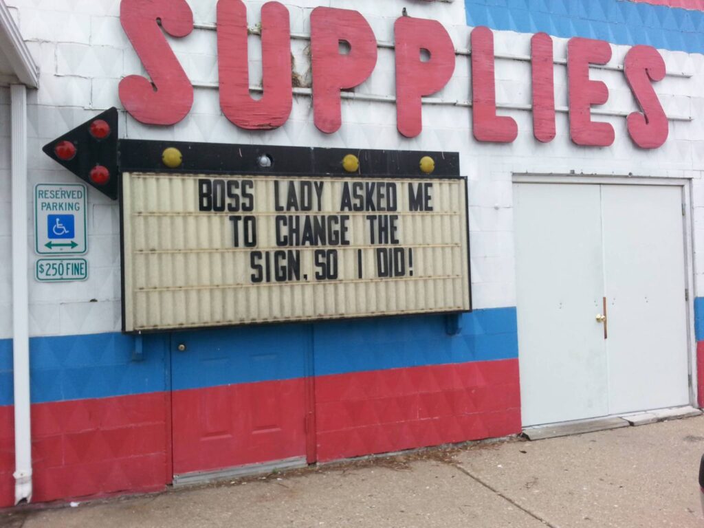 A storefront with the word "SUPPLIES" in large red letters above a marquee sign. The marquee reads, "BOSS LADY ASKED ME TO CHANGE THE SIGN. SO I DID!" A reserved parking sign for handicapped parking with a $250 fine is visible to the left of the marquee. The storefront is painted in red, blue, and white stripes.